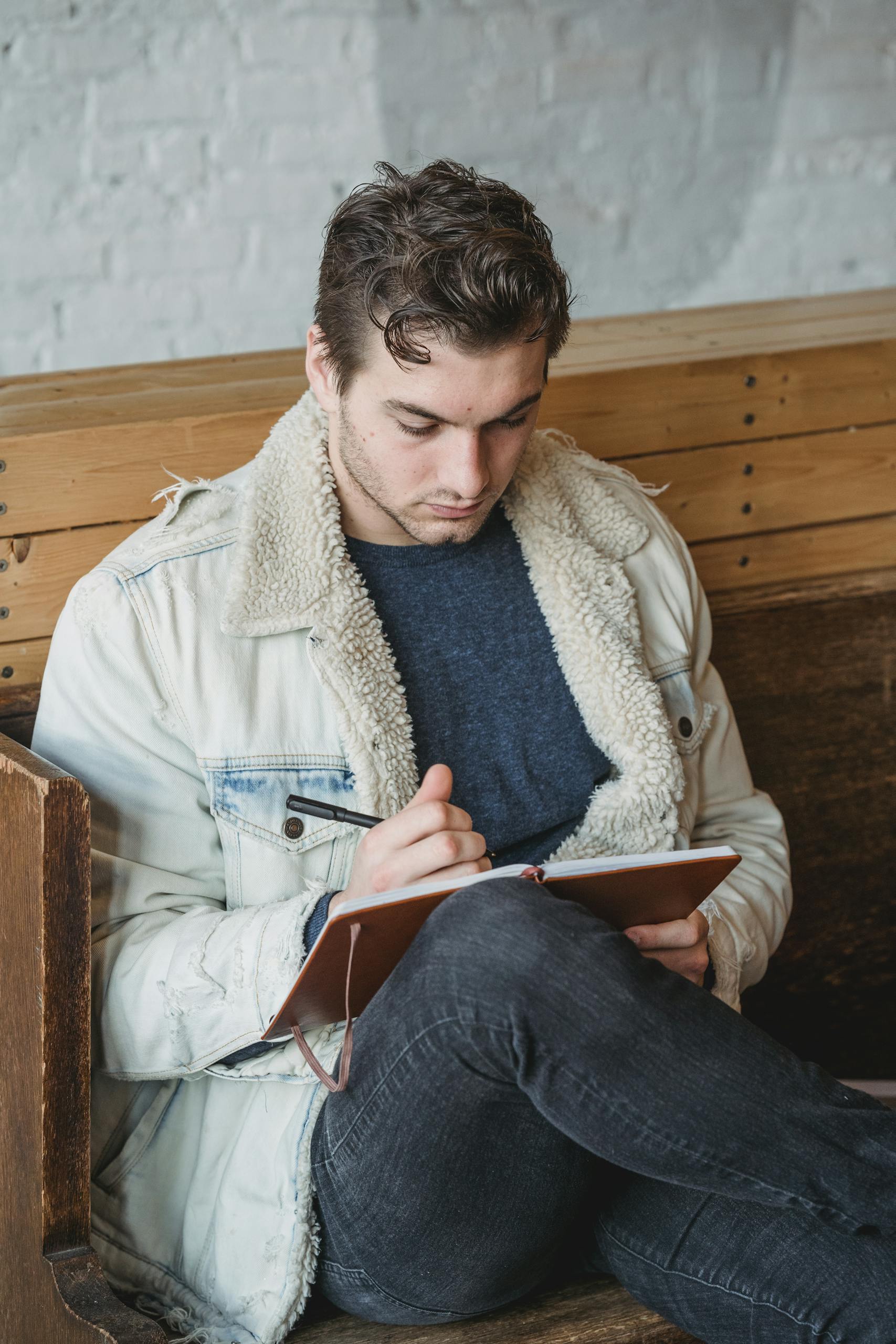Young man intently writing in a notebook while sitting indoors on a wooden bench.